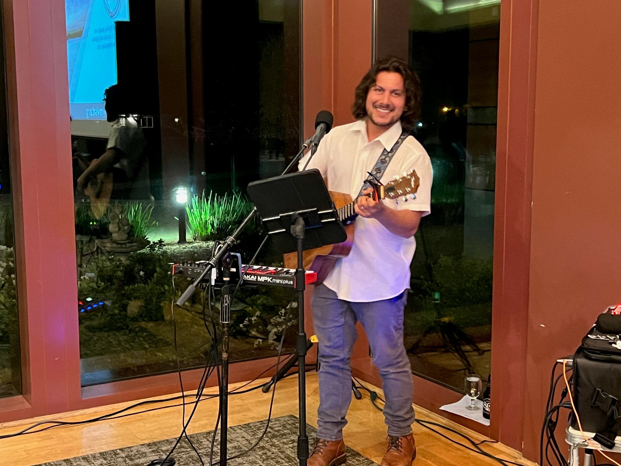 A person playing guitar and singing into a microphone inside a room with large windows.