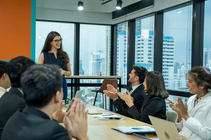 A woman presenting at a meeting with an audience applauding in an office with a city view.