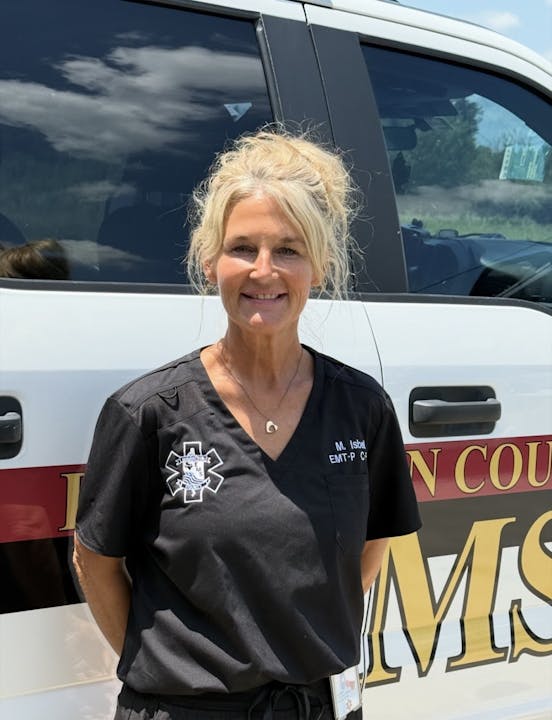 The image shows a smiling woman in a black EMT uniform standing in front of a vehicle with "EMS" visible.