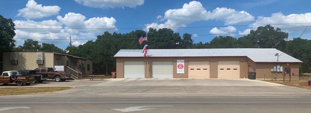 A fire station with three garage doors, the American flag, and a vehicle parked outside under a blue sky with clouds.