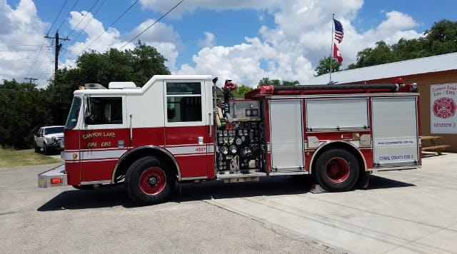 A red and white fire truck parked outside a fire station with an American flag in the background.