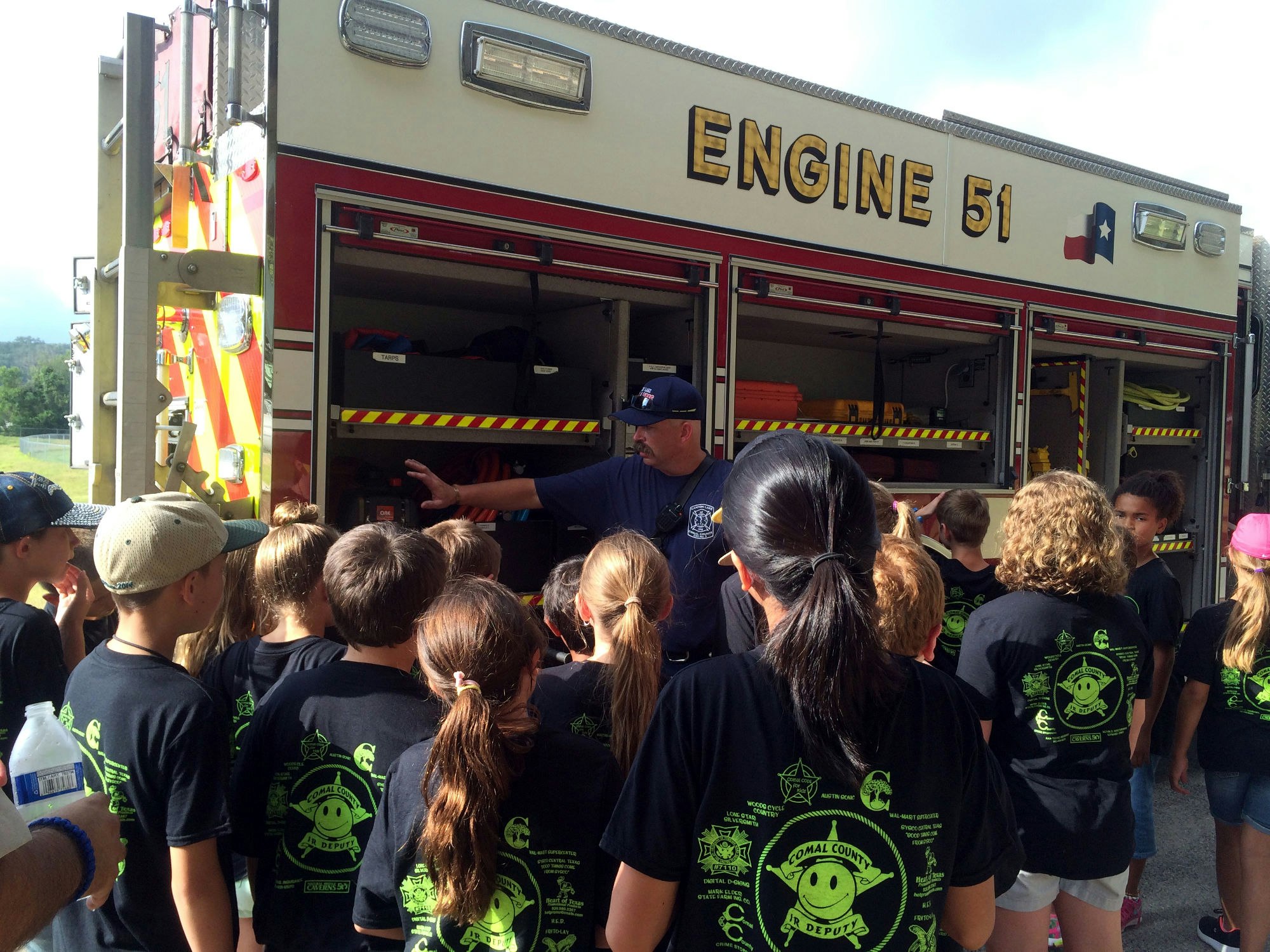 A firefighter is showing equipment to a group of children wearing matching shirts next to a fire engine labeled "ENGINE 51."