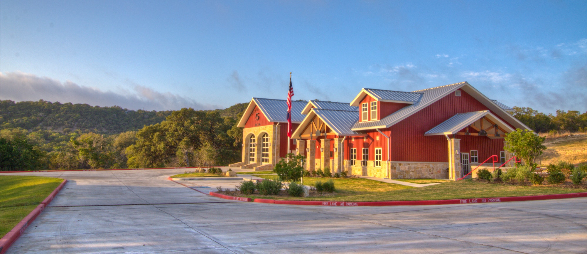 Red and beige fire station with flagpole and parking lot amidst greenery under a clear sky.