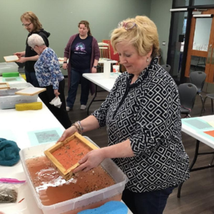 A papermaking workshop with a woman lifting a screen from a pulp-filled basin, and others working in the background.