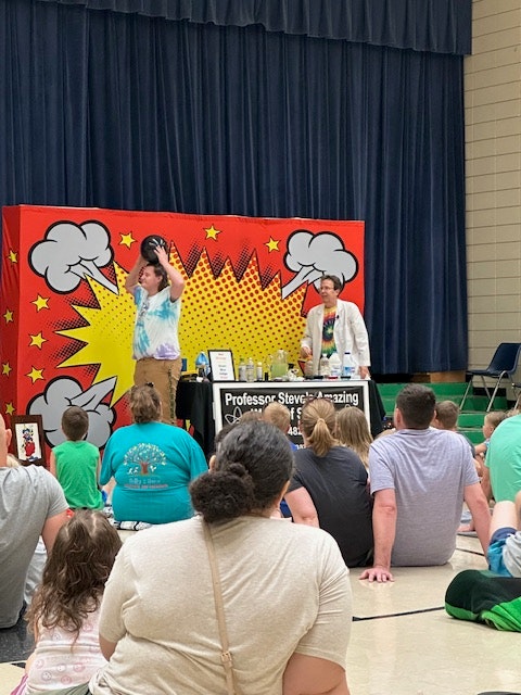 A lively science show with a colorful comic-style backdrop and people watching the demonstration.