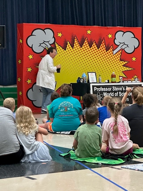 A person in a lab coat performs a science experiment on stage with a colorful backdrop, while children watch from the floor.