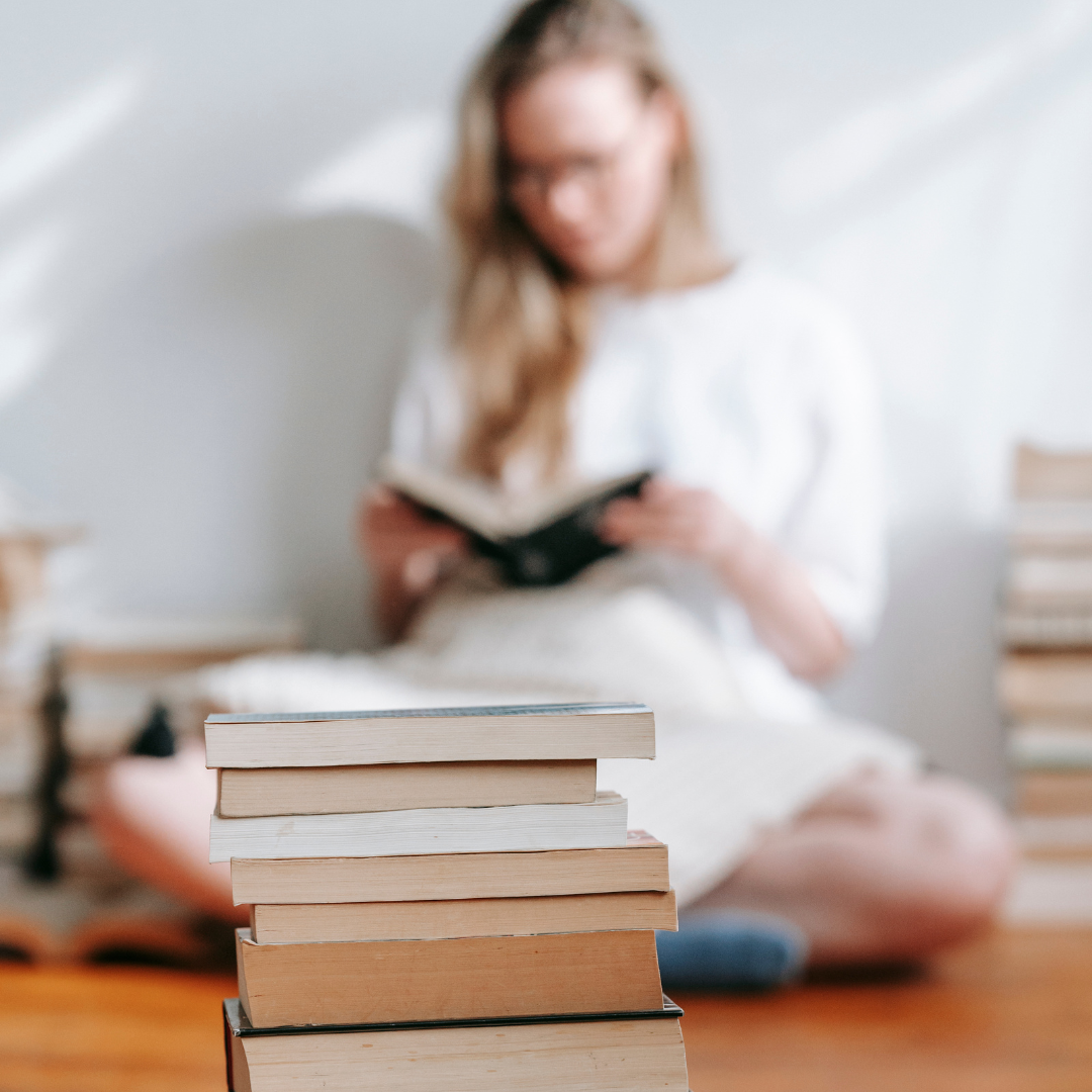 Stack of books in the foreground, with a blurred person reading in the background.