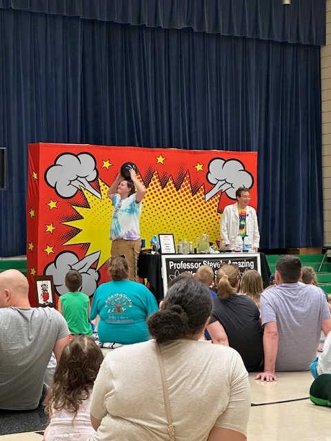 A science show with a colorful comic-style backdrop, presenter on stage, and audience seated on the floor.