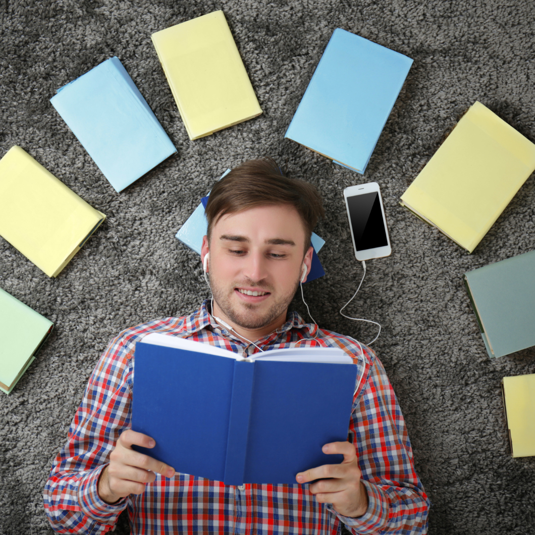 Man with earphones lies among books, holding one open, smartphone beside him.