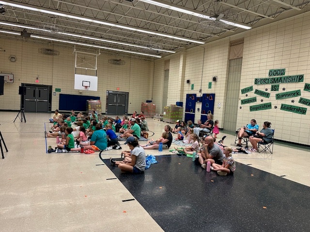 A group of people, mostly children, sitting on a gym floor with sportsmanship posters on the wall.
