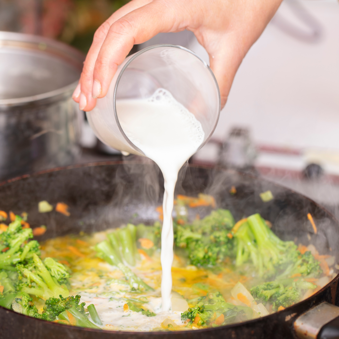 A hand pours milk into a pot with steaming broccoli and vegetables, creating a creamy mixture in the cooking process.