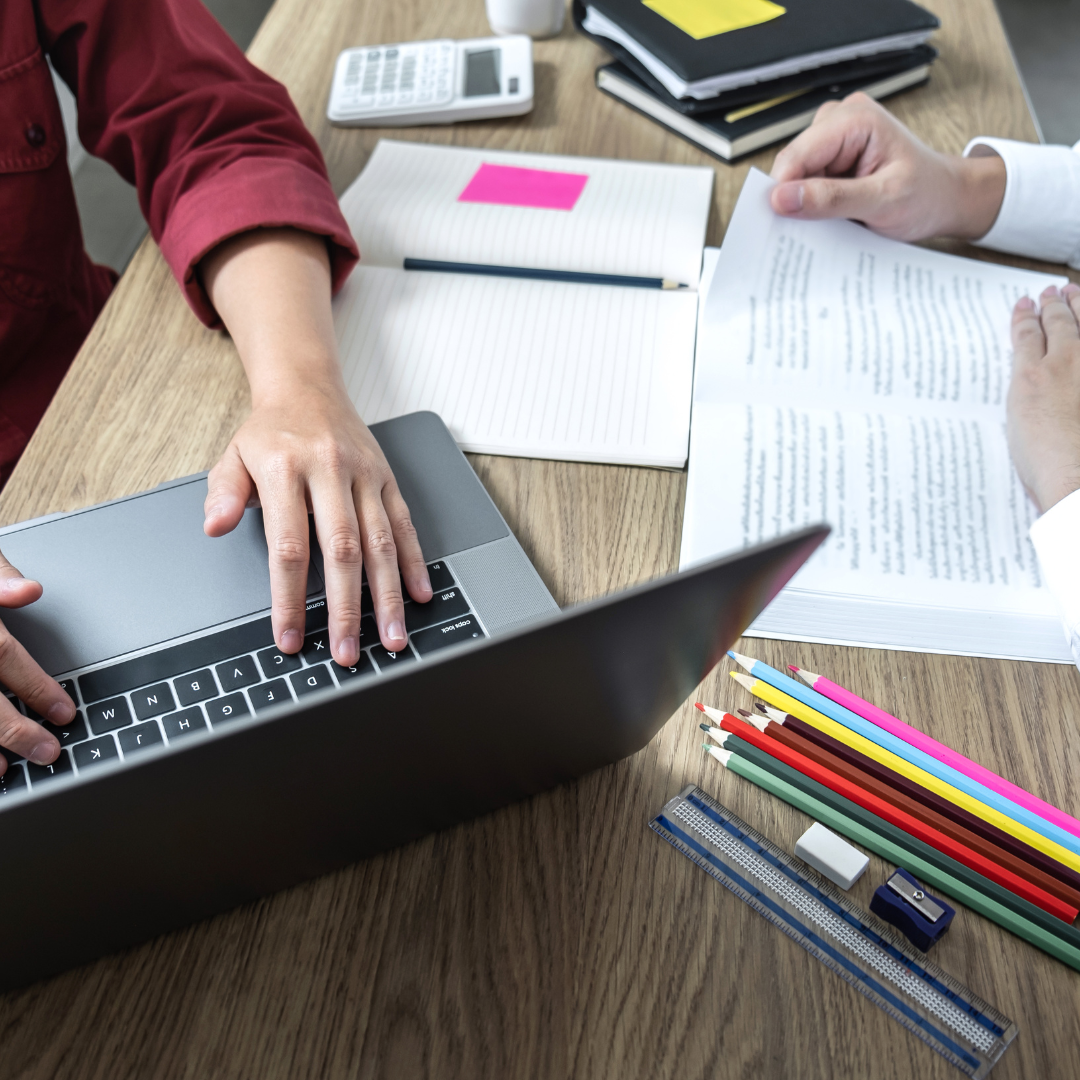 Two people working at a table with a laptop, notebooks, documents, colored pencils, ruler, eraser, and a calculator.