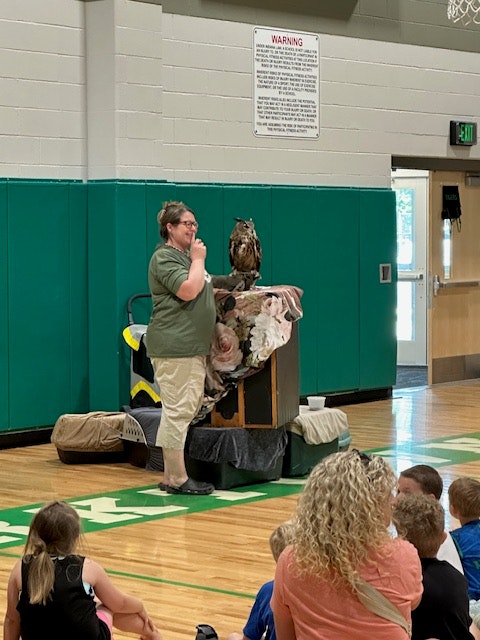 A person speaking to an audience in a gym, with an owl perched nearby.