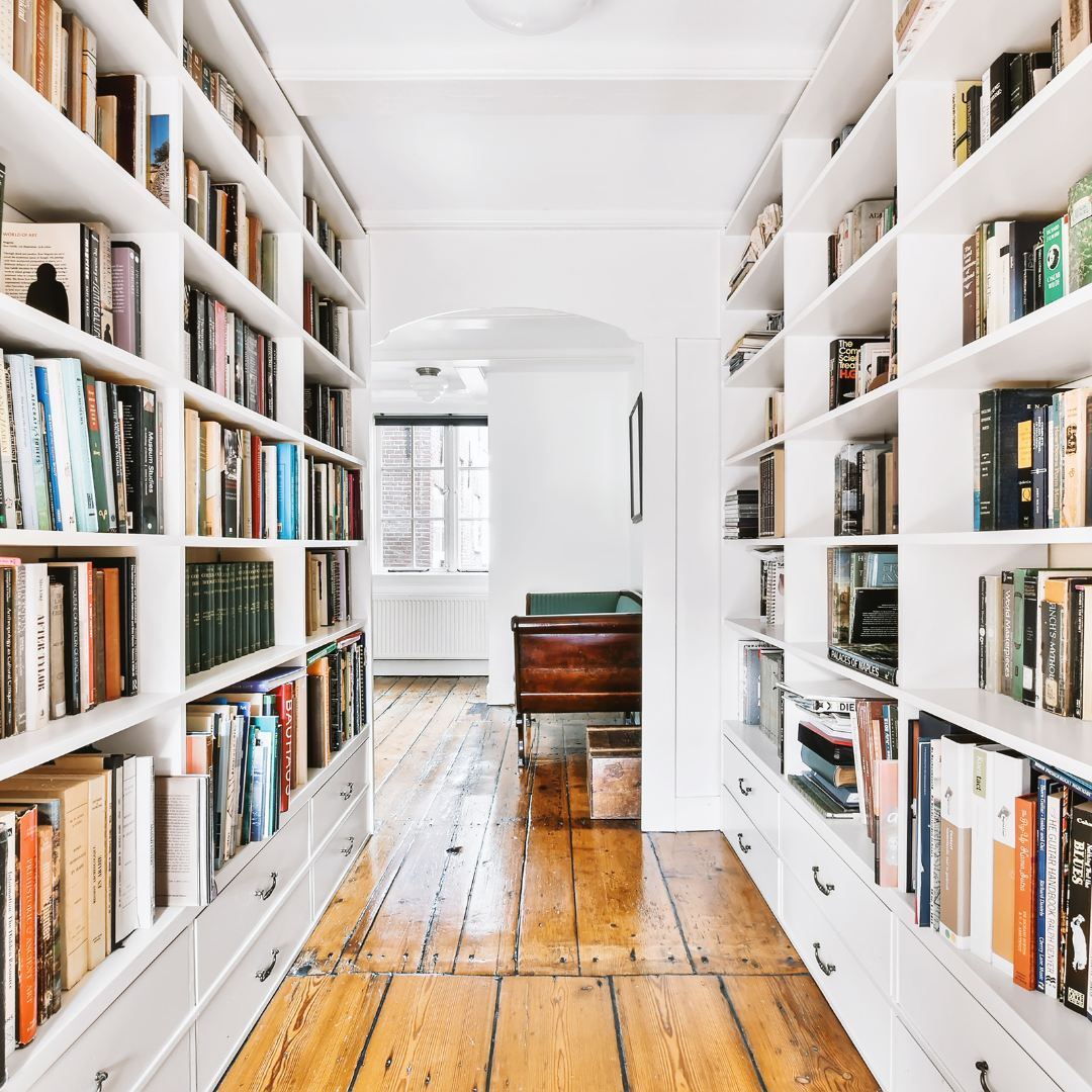 A cozy library corridor with white shelves filled with books, wooden floors, and a glimpse of a bright room with a sofa.