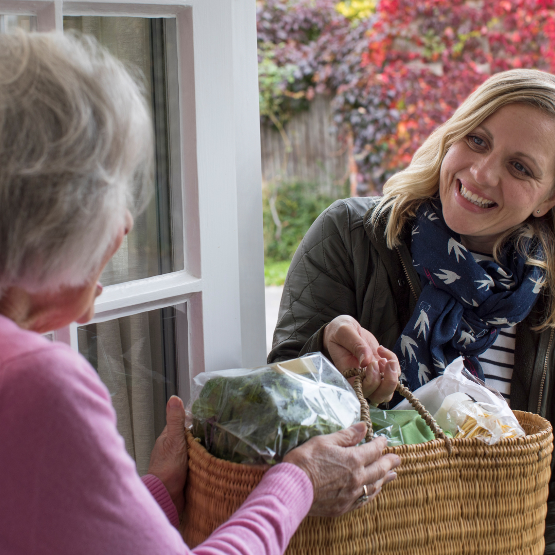 A woman hands a basket of groceries to another at a door.