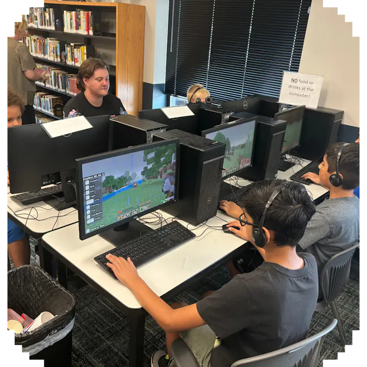 Students with headphones playing a block-style video game on computers in a classroom, with a 'No food or drinks' sign.