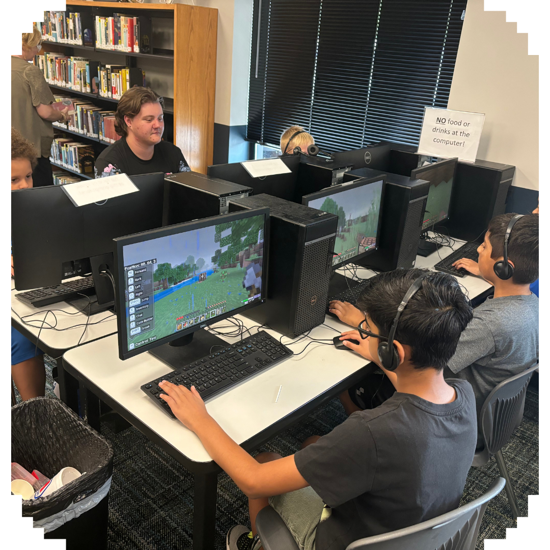 Students with headphones playing a block-style video game on computers in a classroom, with a 'No food or drinks' sign.