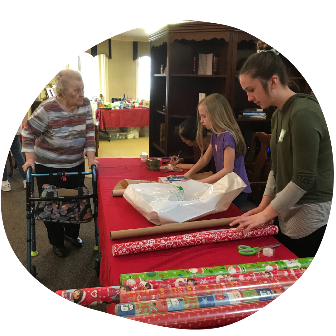 An elderly person with a walker looking at two young people wrapping gifts with colorful paper.
