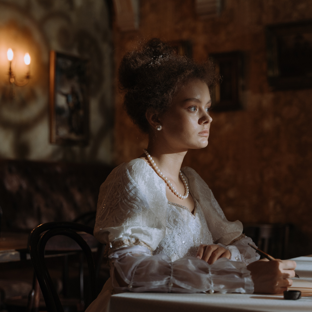 A woman in a vintage white dress and pearl necklace sits at a table, writing, with soft lighting and decorative walls in the background.