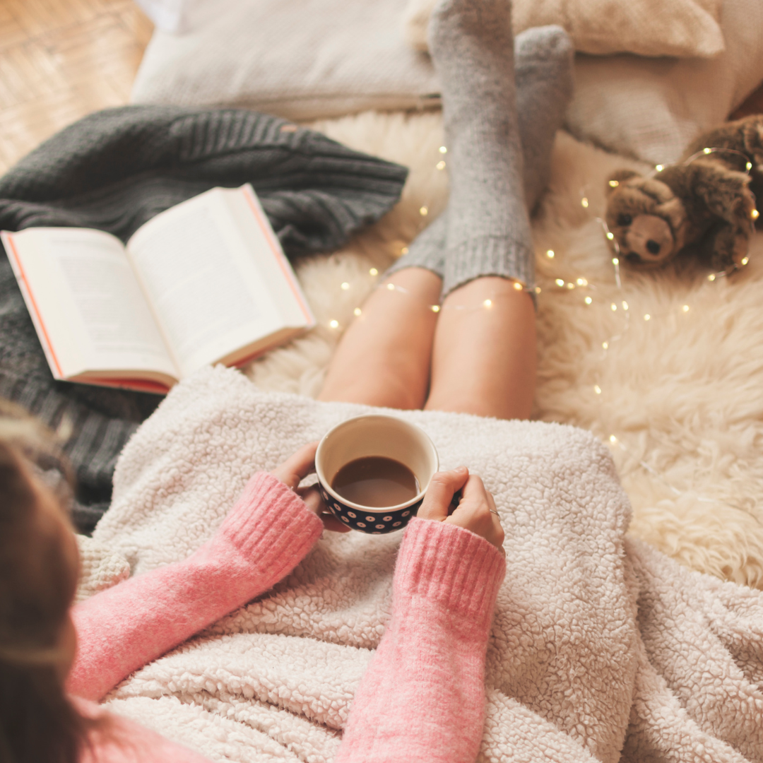 Cozy scene with a person in socks holding coffee, a book nearby, on a fluffy rug with fairy lights.