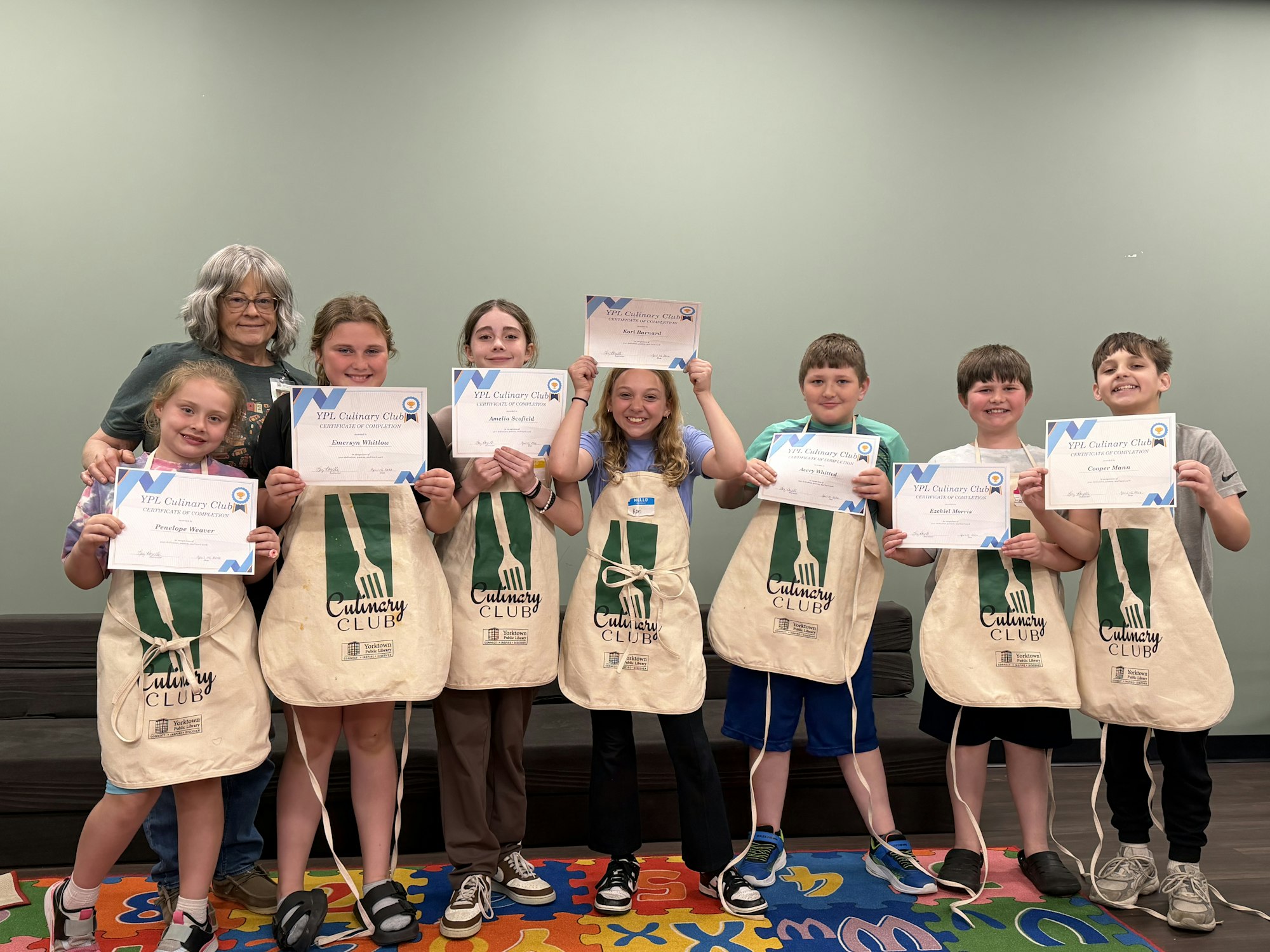 A group of kids poses with certificates at a culinary club, showing their achievements while wearing matching aprons.
