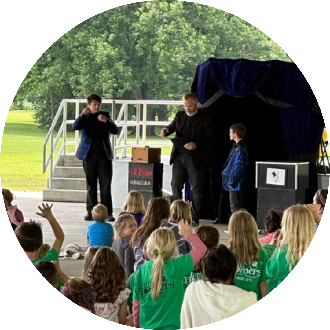A magician performs for a group of children wearing green shirts at an outdoor event.