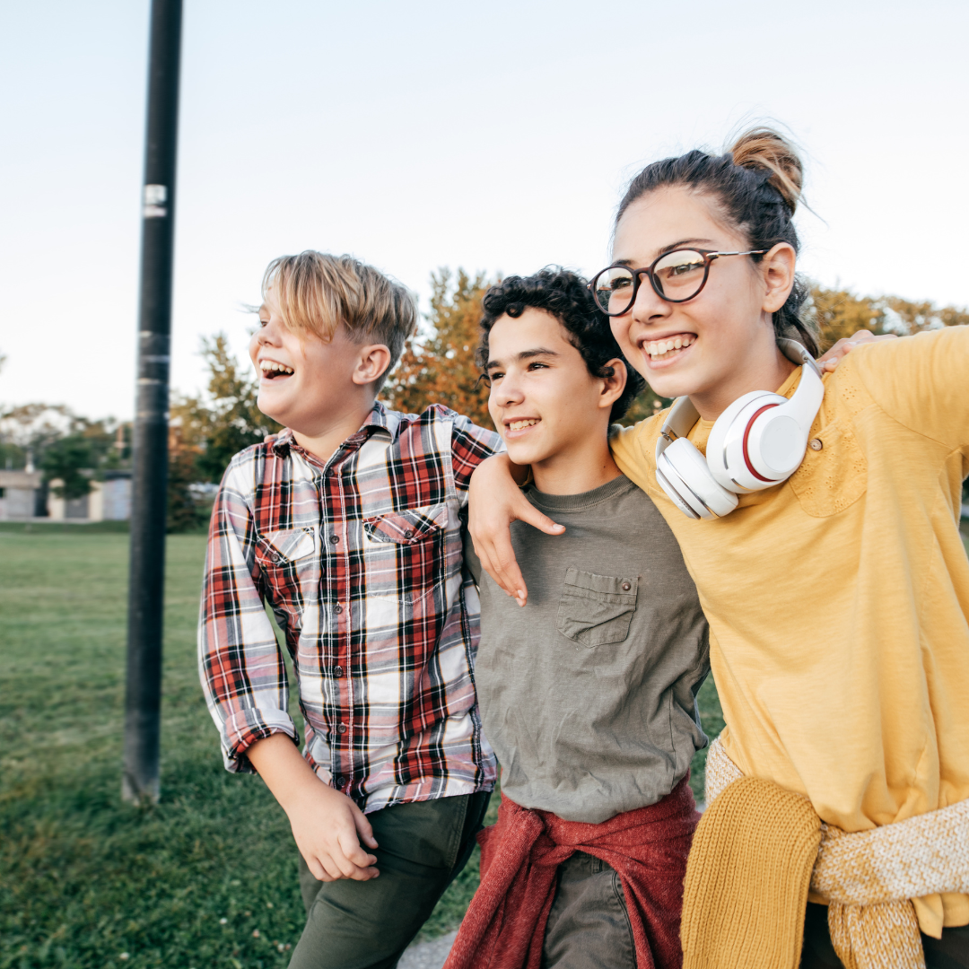 Three smiling kids with arms around each other in a park, one wearing headphones.