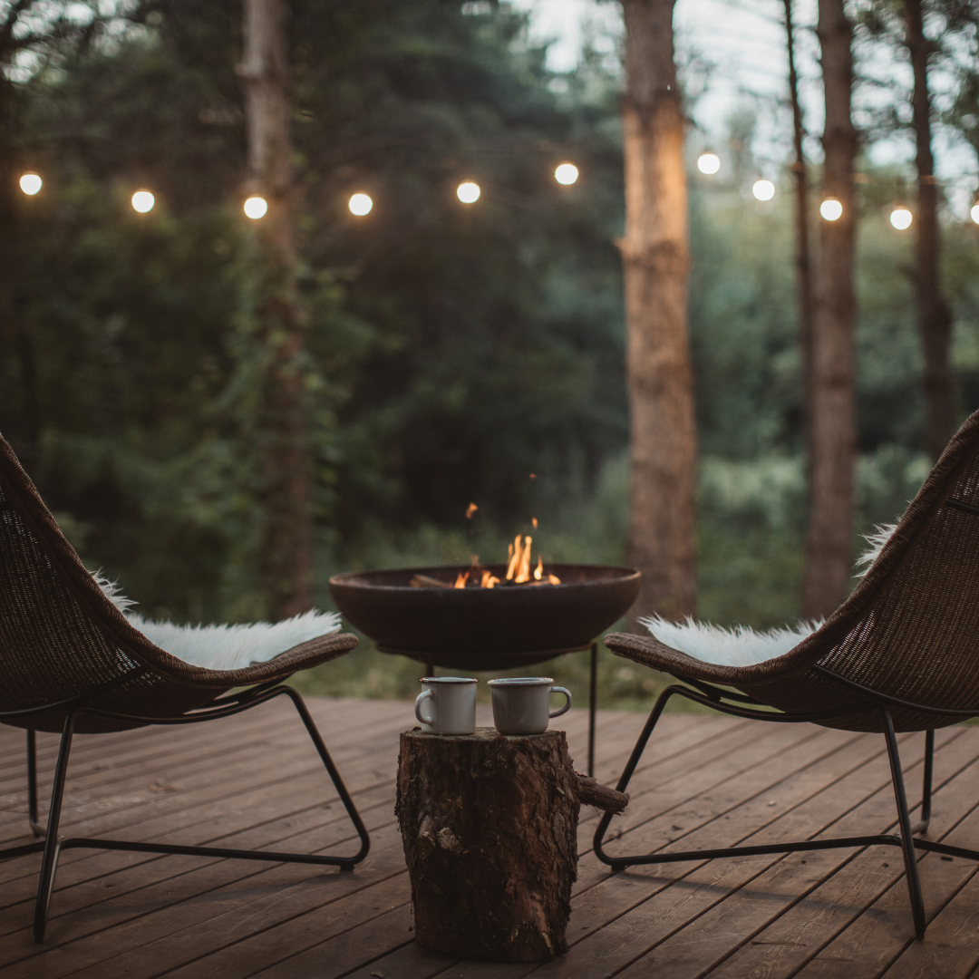 A cozy outdoor setup with two chairs, a fire pit, string lights, and two mugs on a wooden stump.