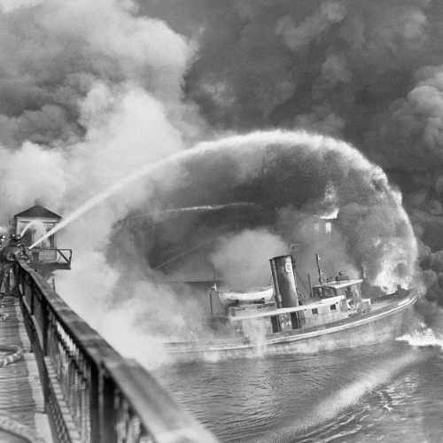 A tugboat sprays water in a smoky scene, possibly battling a fire near a pier, captured in black and white.
