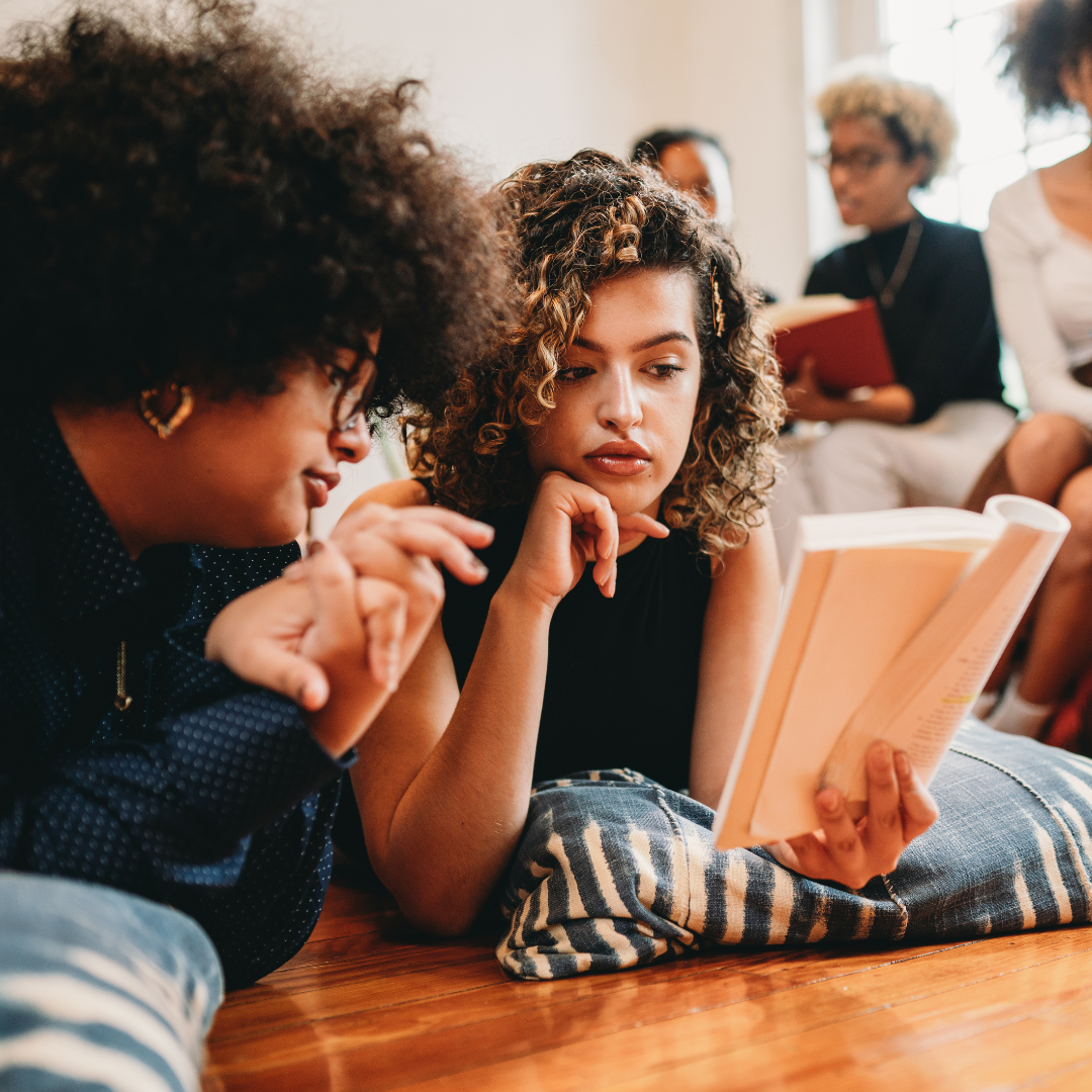 Two people are lying on the floor, reading a book, while others sit in the background with books.