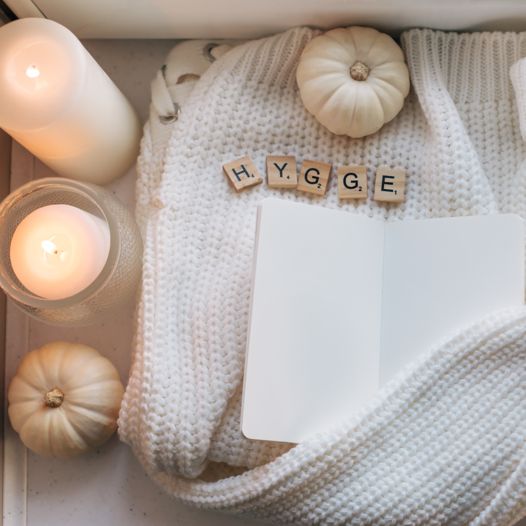 Cozy setting with candles, white pumpkins, a knit blanket, an open notebook, and "HYGGE" spelled in tiles.