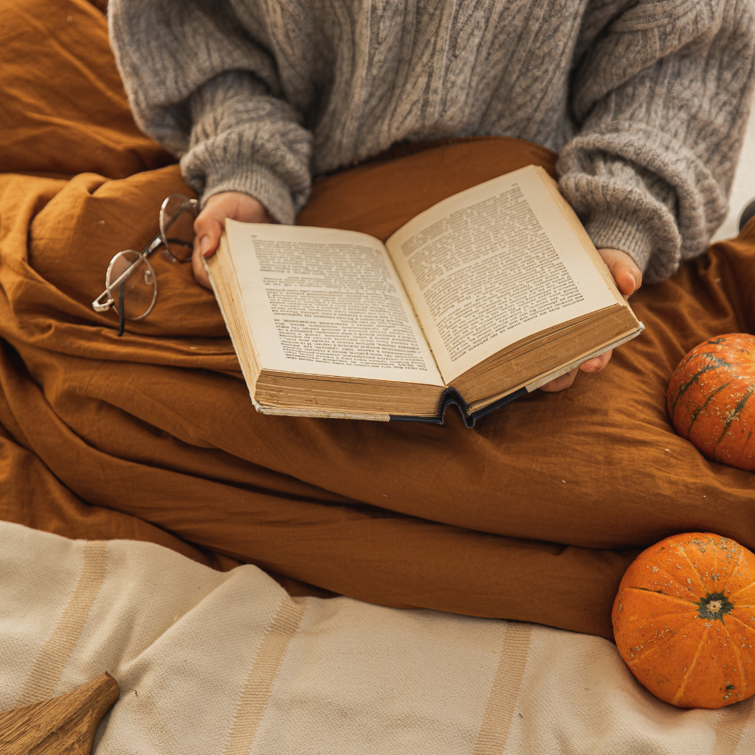 A person in a cozy sweater reads a book on a warm blanket, accompanied by glasses and small pumpkins.