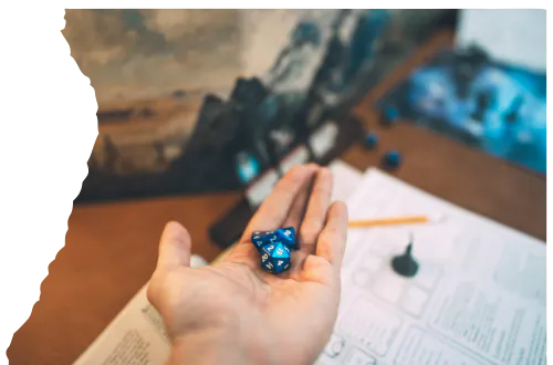 A hand holding a blue dice, with a tabletop gaming setup, including game rules, a pencil, and a mini figure in the background.