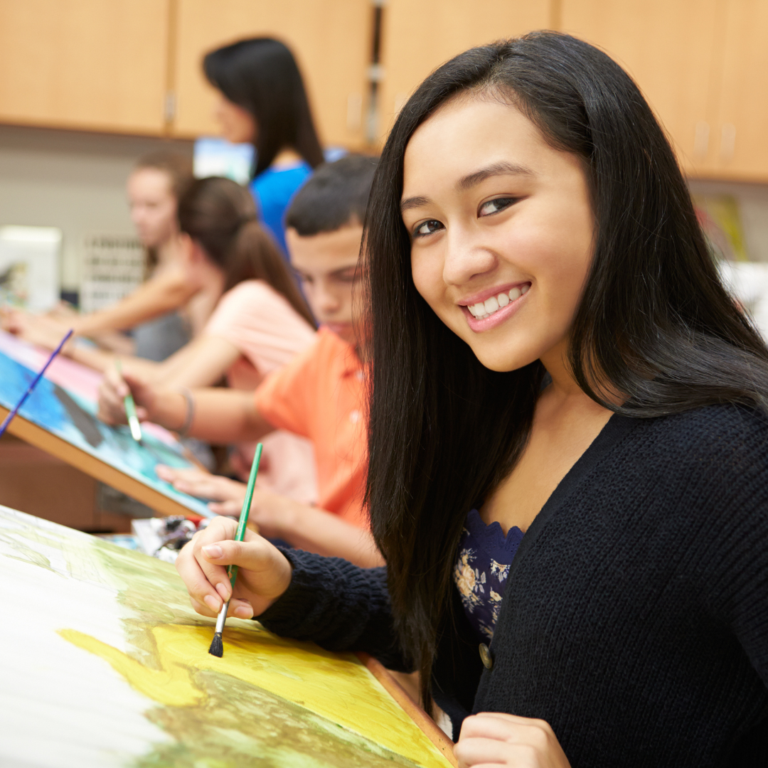 A group of students engaged in a painting class, with a smiling girl in the foreground.