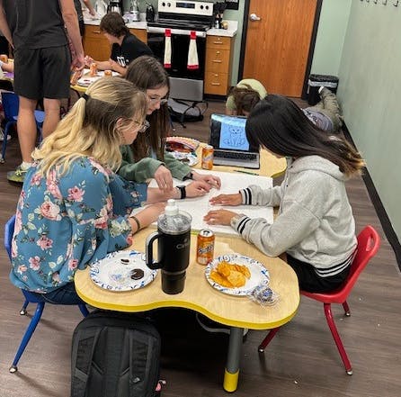 A group of people in a room, sitting at tables with snacks and drinks, working on papers, with a laptop open in the background.
