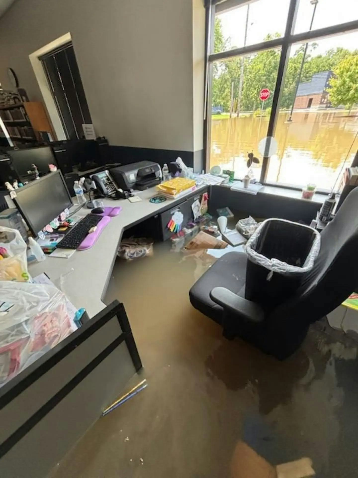 An office space is flooded, with water covering the floor and items scattered around. A computer and chair are also visible.