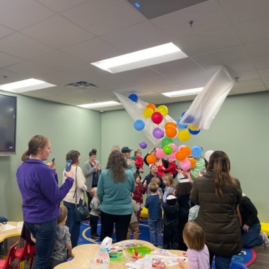 A group of people, including children, gathers to watch colorful balloons being released indoors at an event.