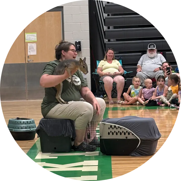 A person holds a small animal while an audience of children and adults watches in a gym setting.