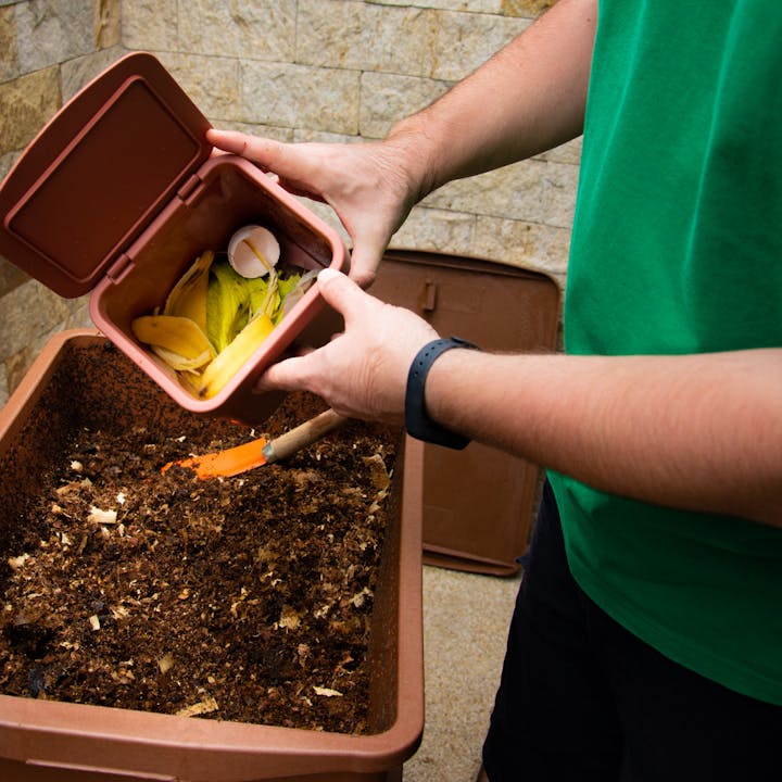 A person is holding a container with food scraps, like banana peels and eggshells, over a compost bin.