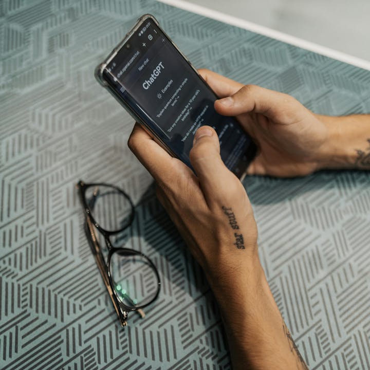 A person is using a smartphone displaying ChatGPT while glasses are resting on a textured surface nearby.