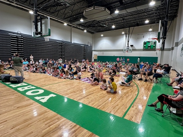 A gym with many seated people and children watching a presentation or event.