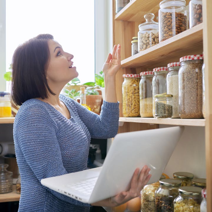 A woman is using a laptop while examining jars of food on a shelf in a bright kitchen.