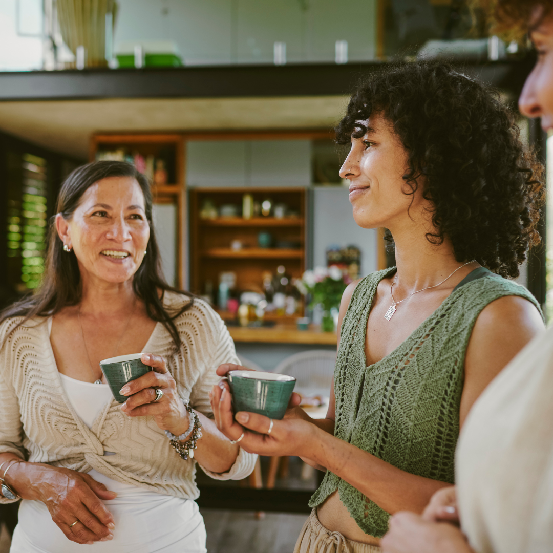 Three women are engaged in conversation, holding small cups, in a warm and inviting indoor space.