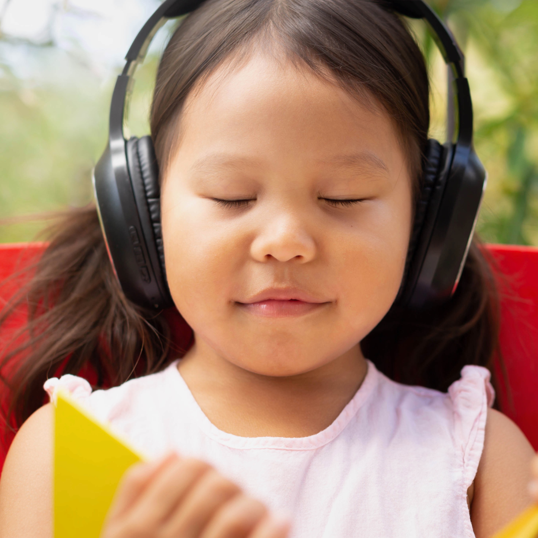 A young child wearing headphones and reading a book, with a serene expression on her face.