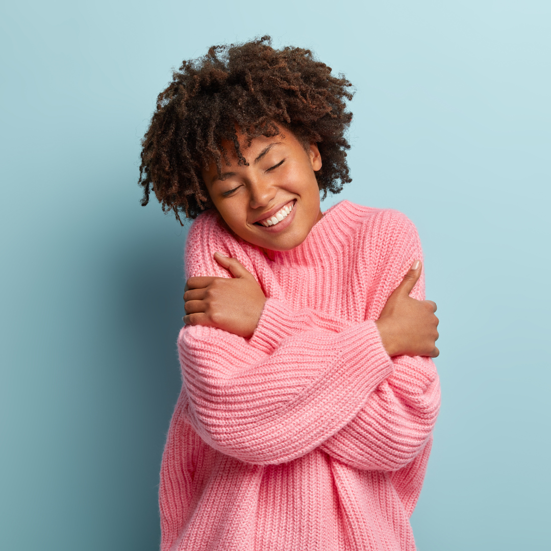 A person smiling with eyes closed, hugging themselves while wearing a pink sweater against a light blue background.