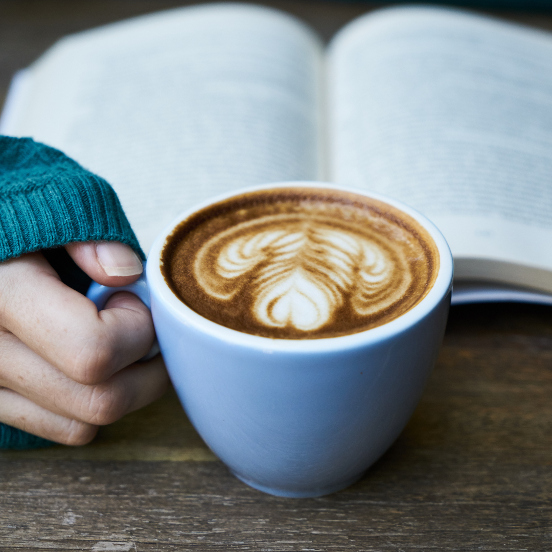 A hand holding a cup of coffee with latte art, next to an open book on a wooden surface.
