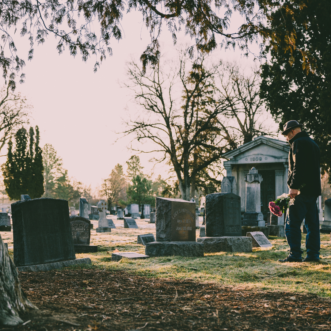 A person holding flowers stands by a gravestone in a cemetery at sunset.