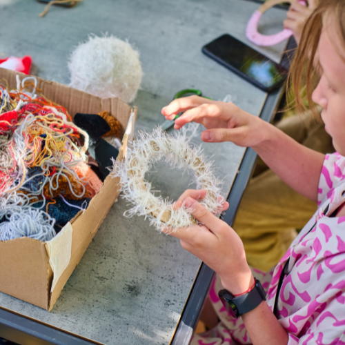 A child holds a fluffy, circular craft made from yarn, surrounded by colorful yarn scraps in a cardboard box on a table.