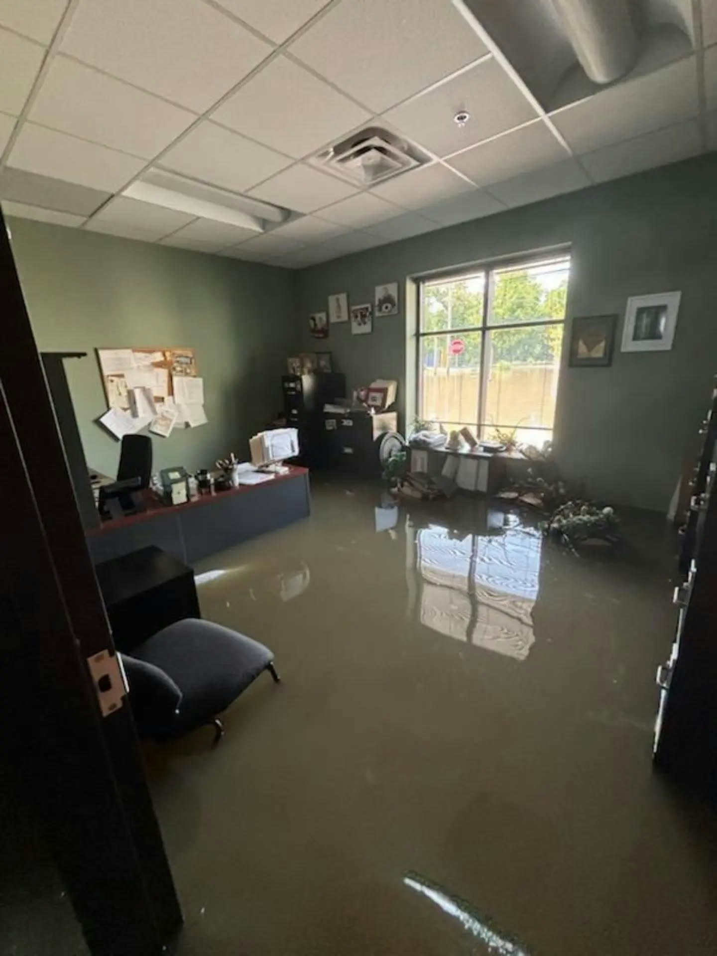 A flooded office with water covering the floor, affecting furniture and workspaces. The environment looks chaotic and damaged.