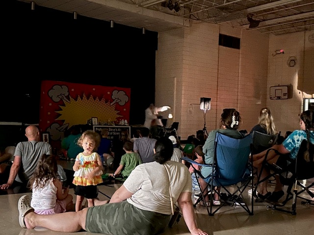 People sitting in a gym watching a performance with a colorful backdrop. A child stands in the foreground.